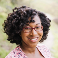 Asya Spears stands outdoors with greenery in the background. She wear eyeglasses and a patterned pink top.