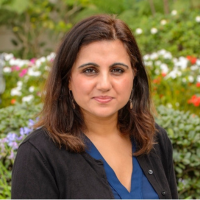 Dr. Ridhima Iyer stands outside against a backdrop of white and red flowers wearing a black blazer and a dark blue top. She has brown shoulder-length hair