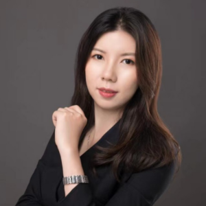 EMPH Student Daisy Wang stands indoors against a grey wall as she has her arm and hand up near her face. She wears a black top and silver watch