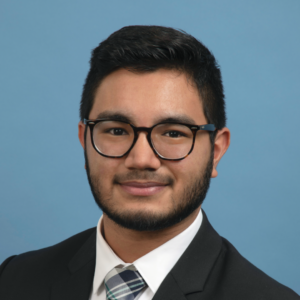 EMPH Student Elías Rodríguez wearing eyeglasses and a suit jacket, white collared shirt, and a tie, standing against a blue background indoors