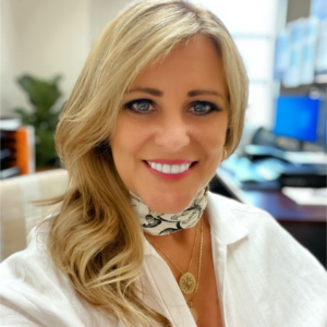 close-up image of EMPH student Lisa Edwards wearing a white colored top, her blond hair over one shoulder as she sits indoors at a desk