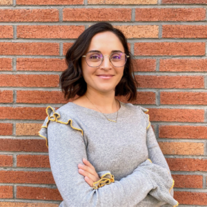 EMPH Student Maite Medina stands outdoors with her arms crossed in front of her as she stands against a red brick wall. She wears a light grey sweater and glasses