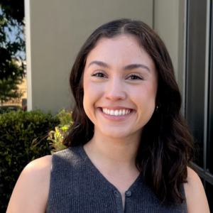 EMPH Student Maya Camacho standing outside with a green hedge behind her and a white building wall